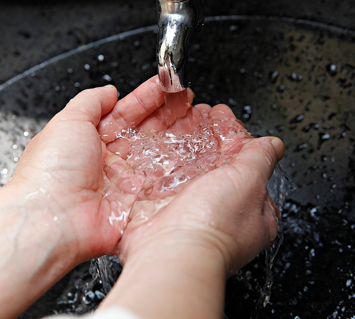 Hands cupped under running water from a faucet over a black sink.