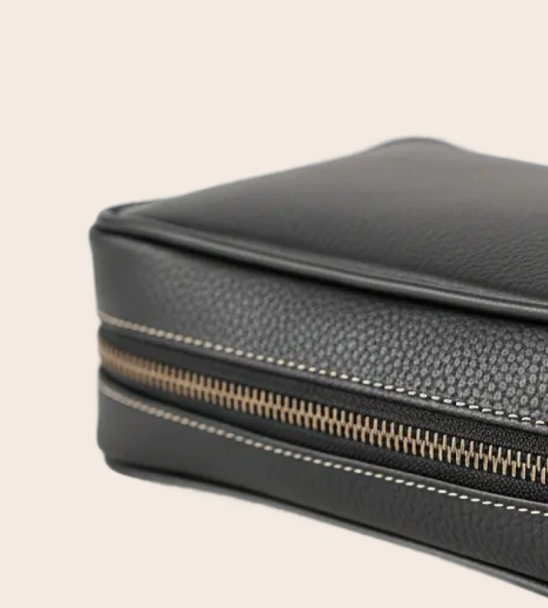 Close-up of a black leather bag corner showing texture, stitching, and metal zipper details.