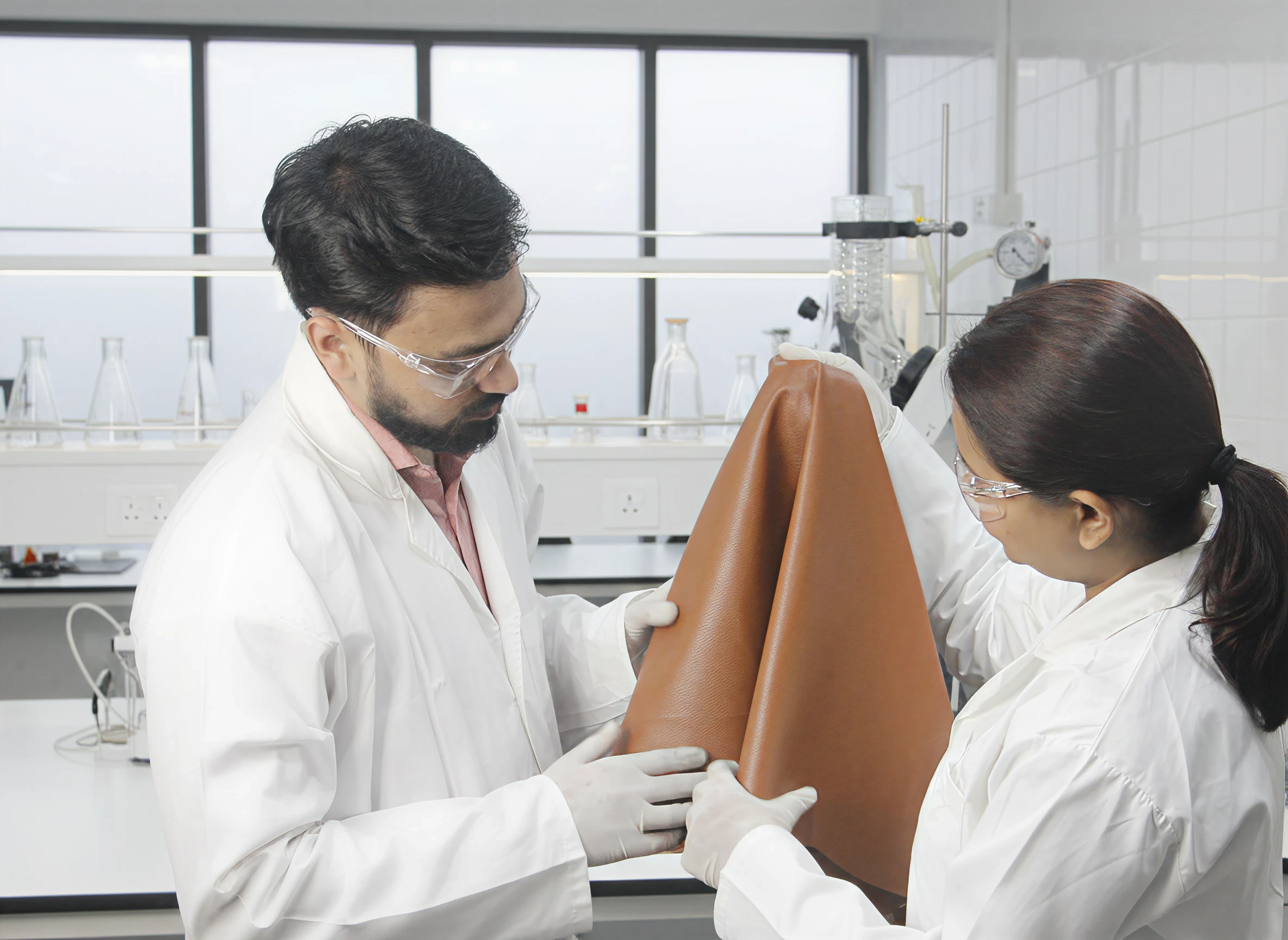 Two scientists in lab coats inspecting a brown leather sheet in a laboratory setting.