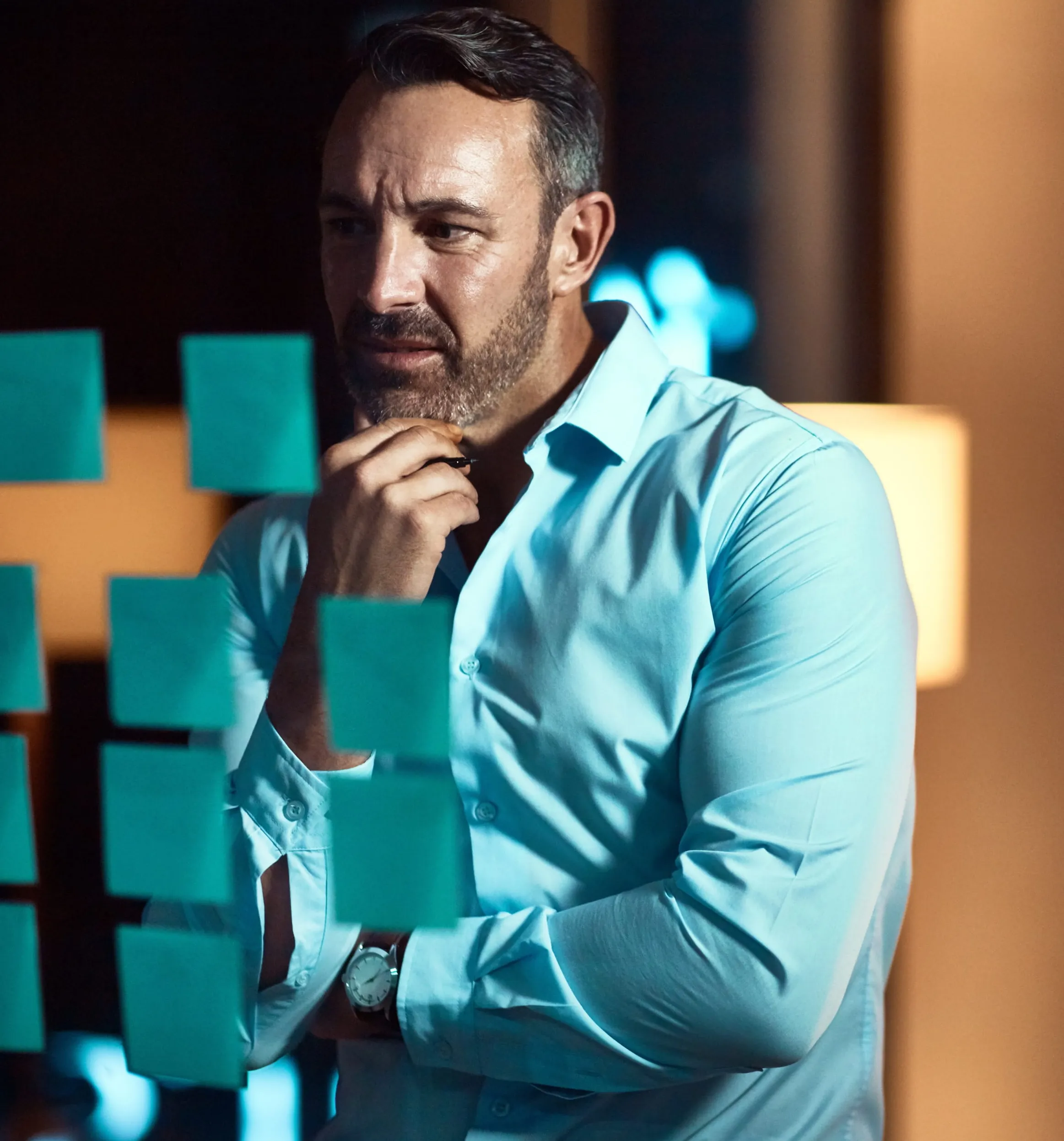 Thoughtful man in a light blue shirt looks at blue sticky notes on glass in a dimly lit room.