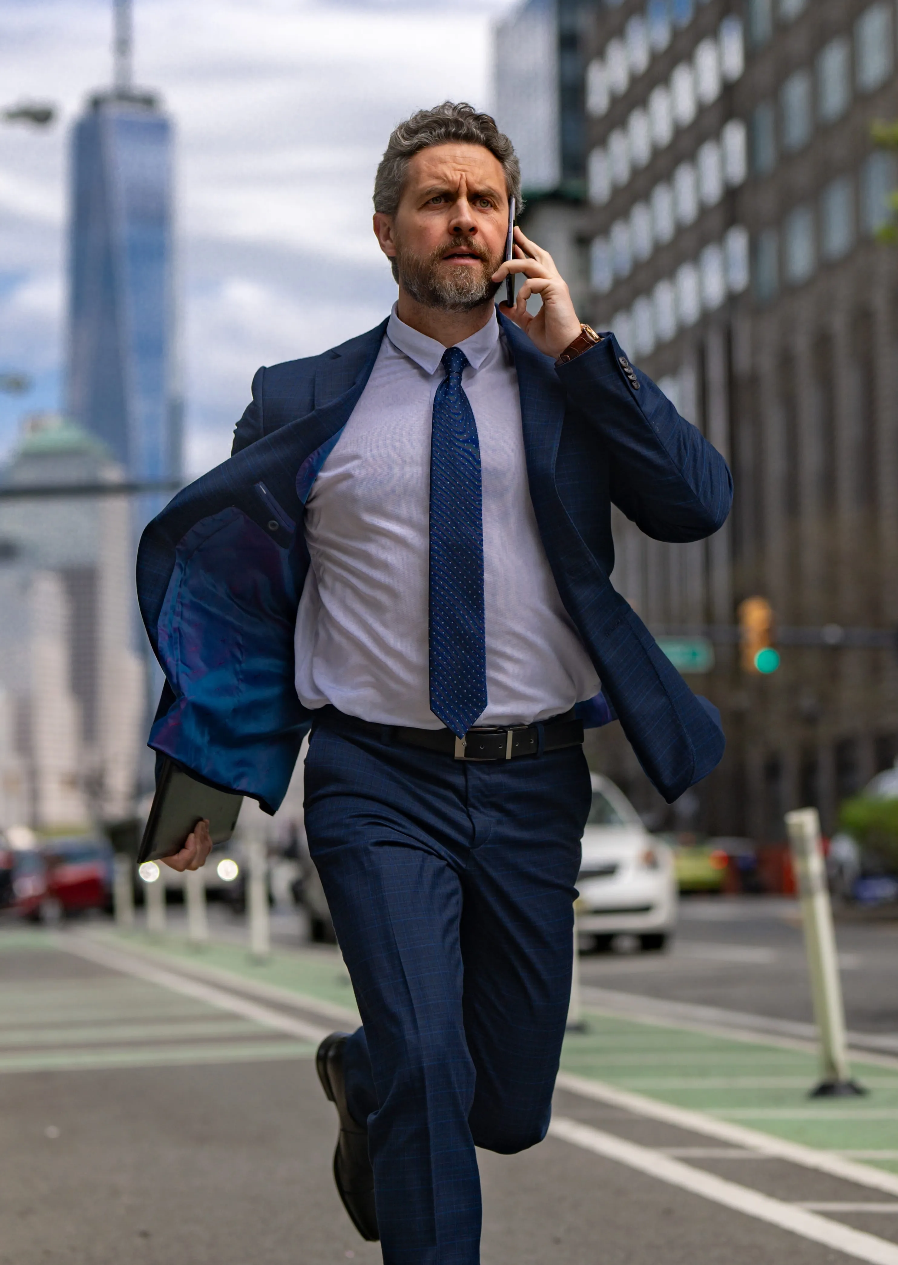 Man in a blue suit running on a city street while talking on a smartphone and holding a tablet.
