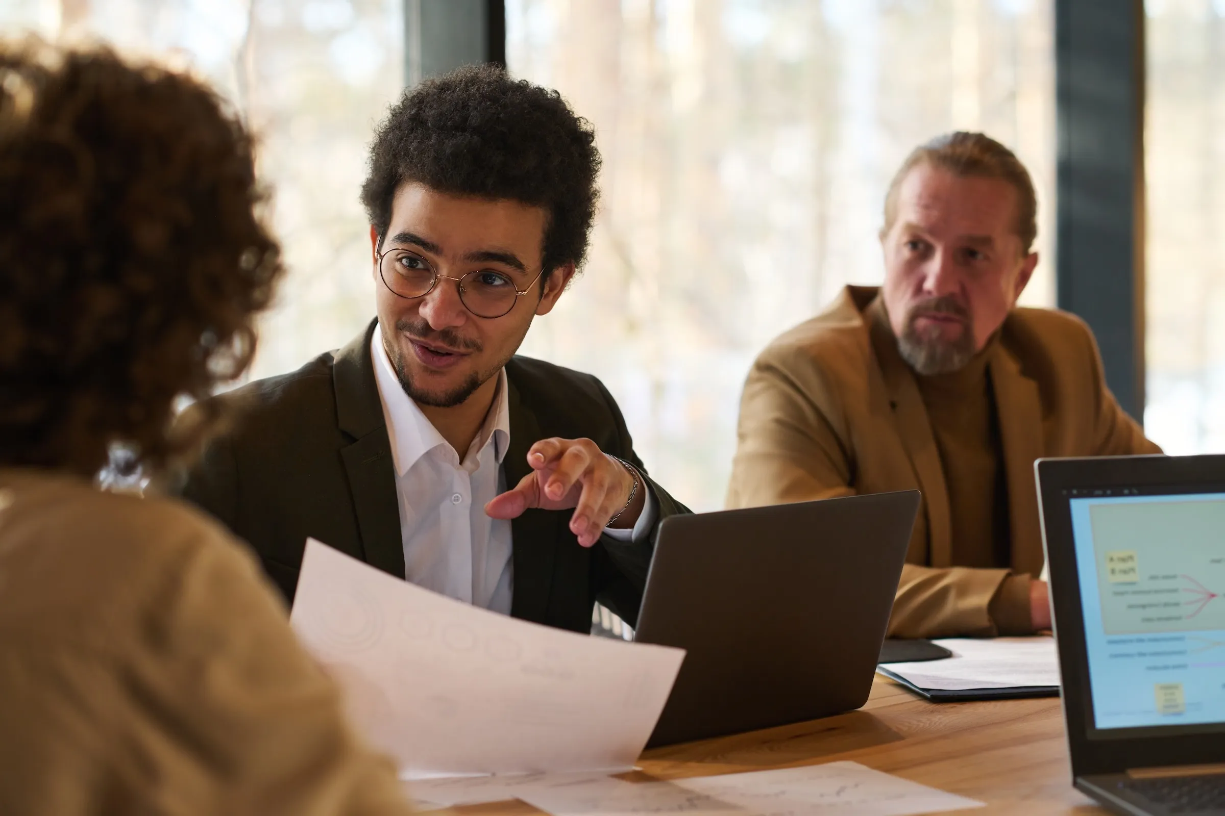Businessman with glasses explaining a document to a colleague while another man watches in a meeting room.