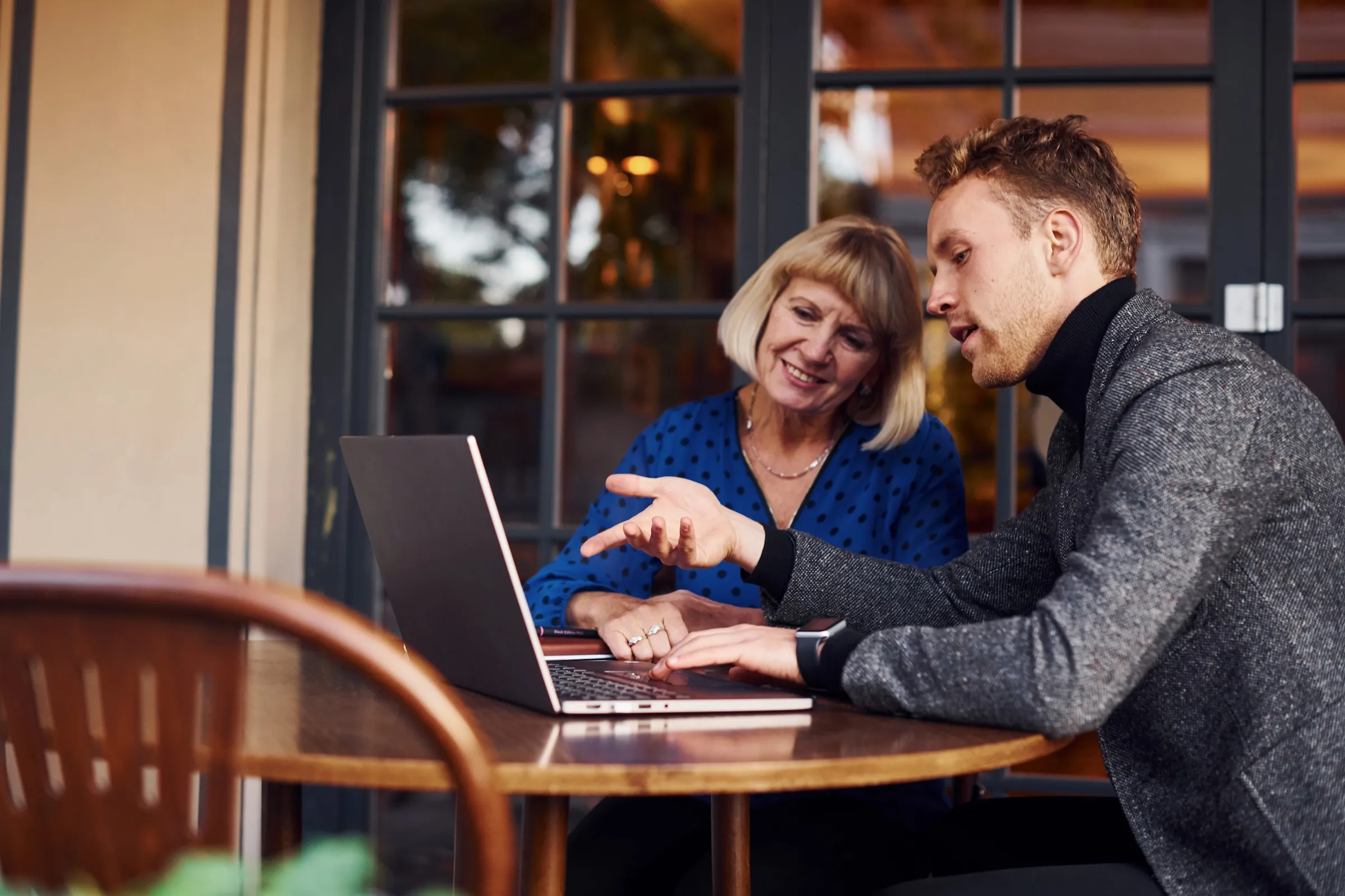 A young man and an older woman sitting at a table, discussing something while looking at a laptop screen.