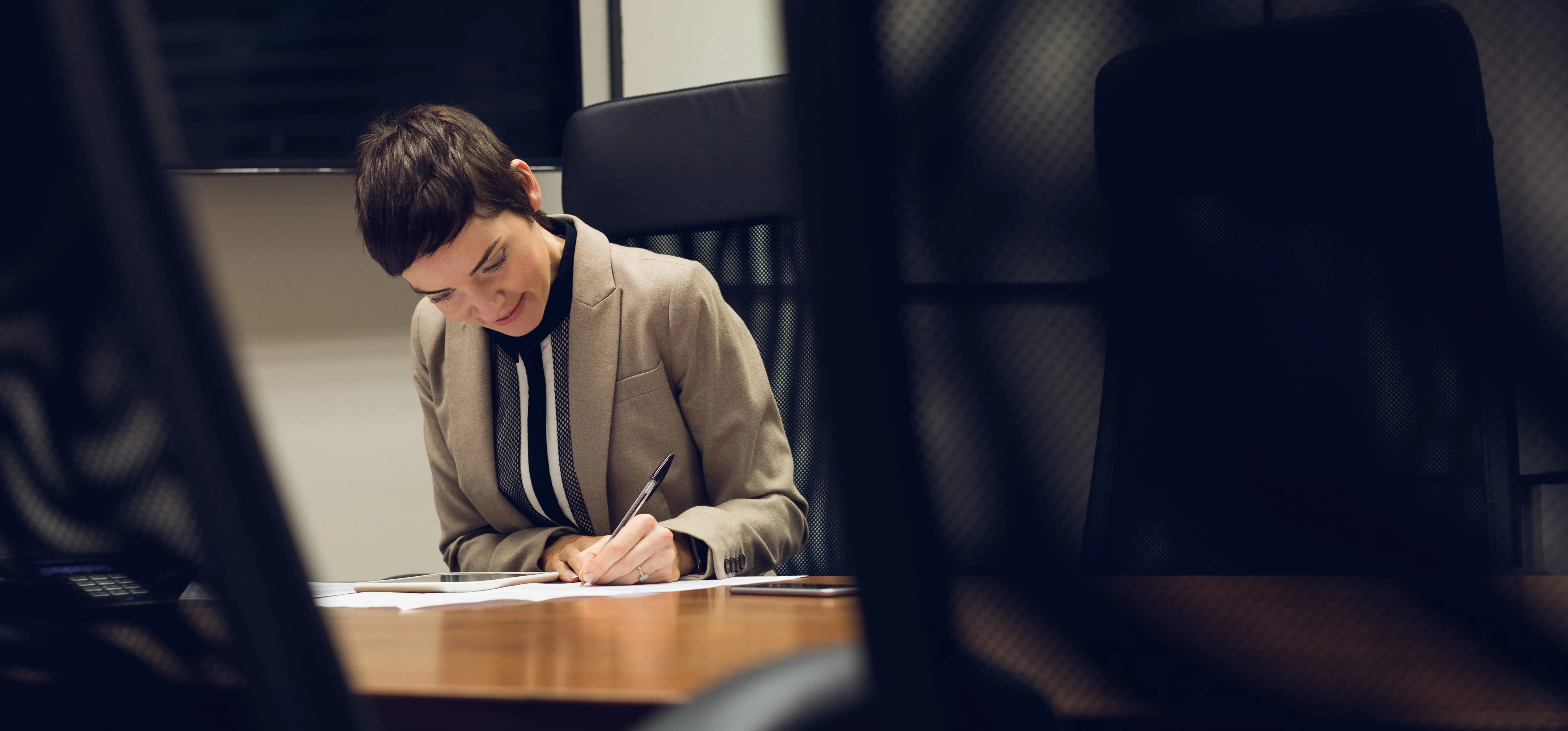 Businesswoman with short hair sitting at a desk, writing on paper with a pen in a modern office setting.