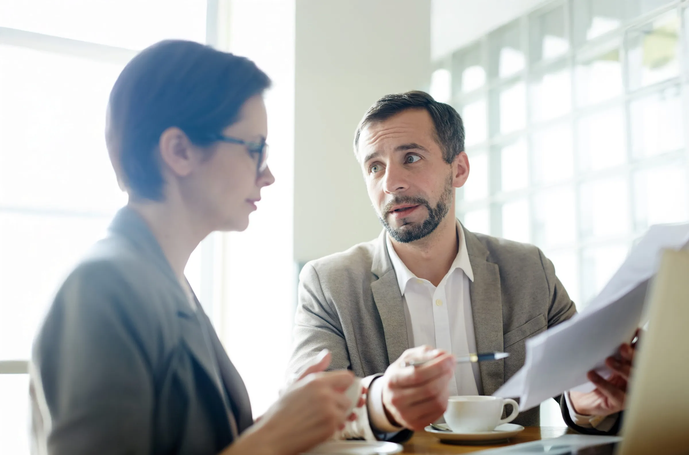 Man in beige jacket discussing documents with woman wearing glasses over coffee in a bright office.