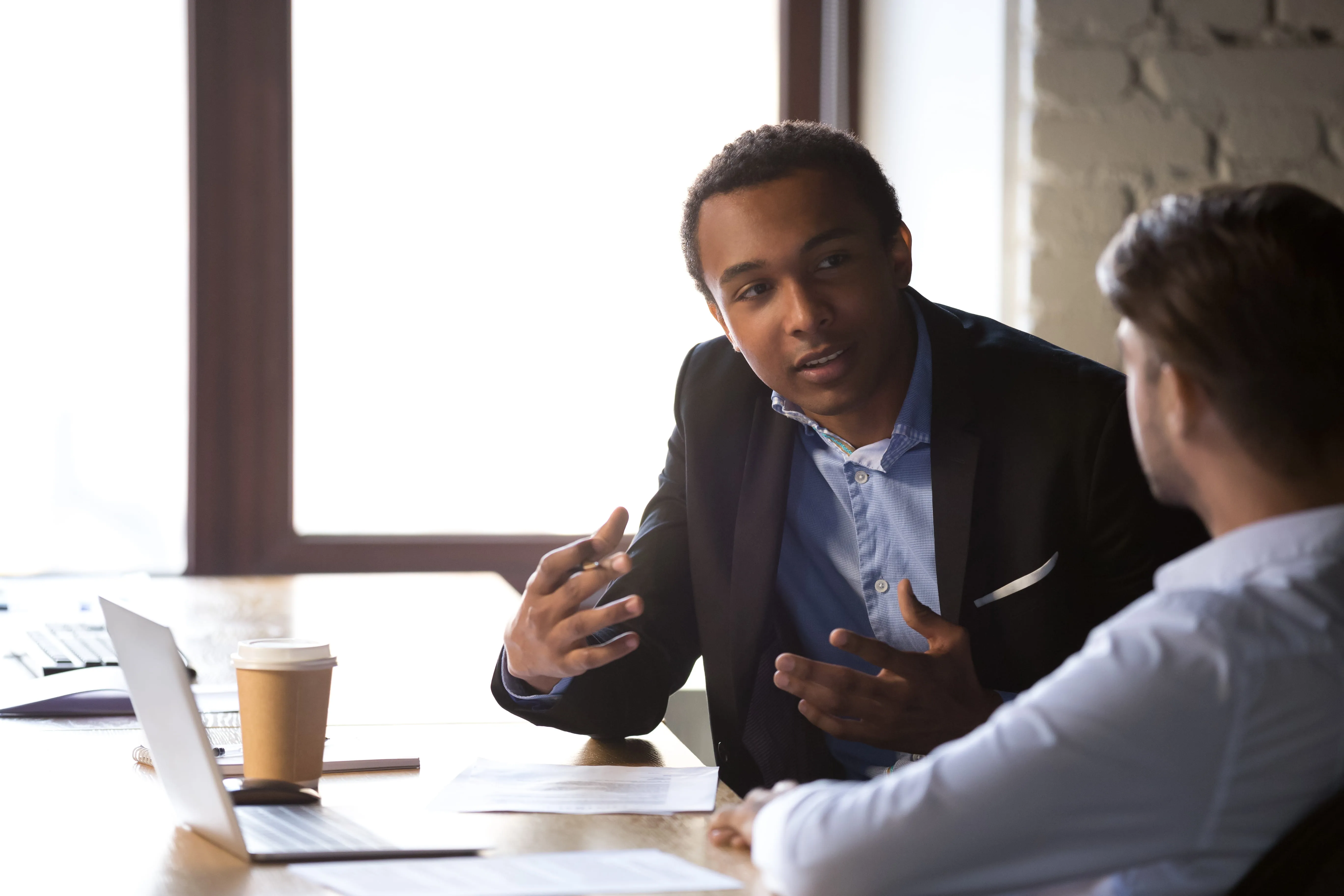 Two businessmen having a discussion at a table with a laptop and coffee cup near a window.