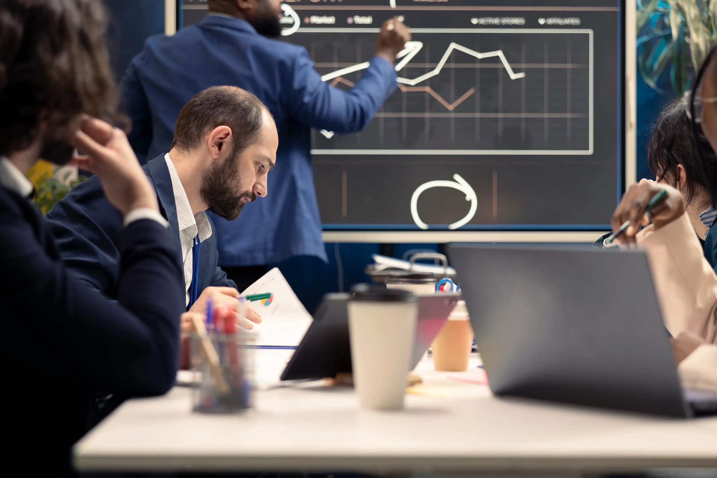 Business professionals in a meeting room reviewing charts on a large screen and documents, with laptops and coffee cups on the table.