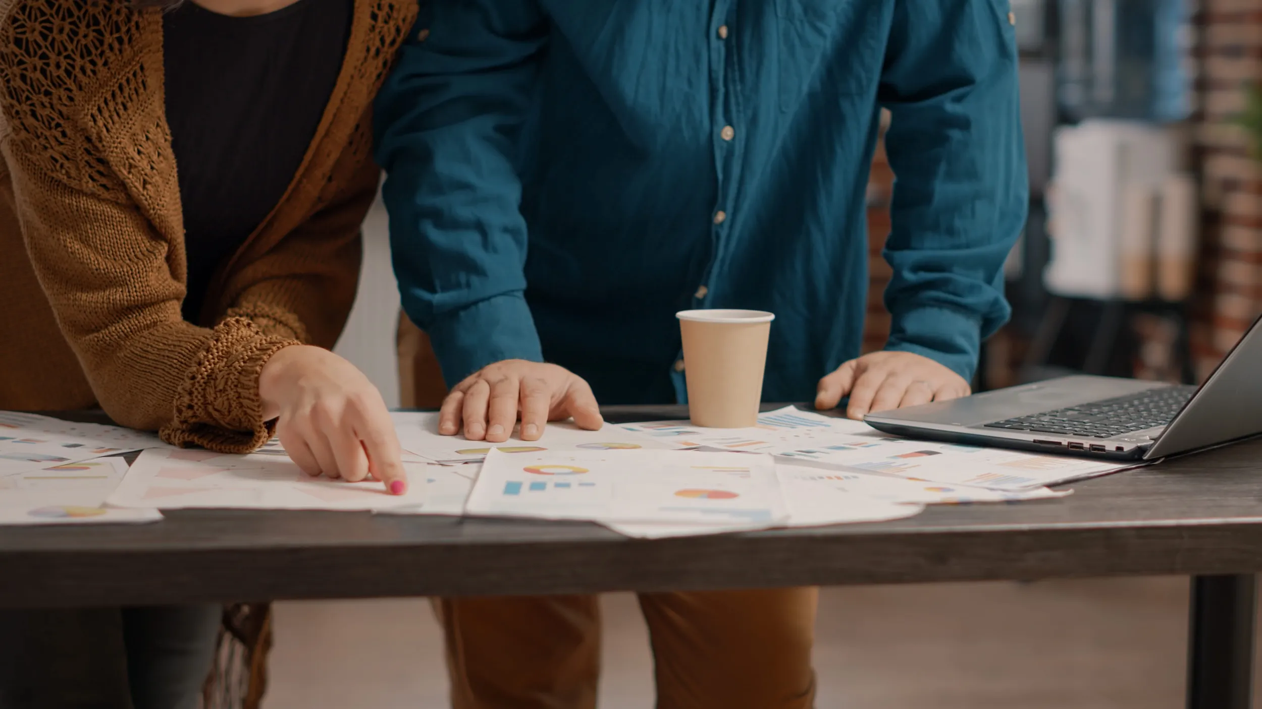 Two people reviewing printed charts and graphs on a table with a laptop and a disposable coffee cup.