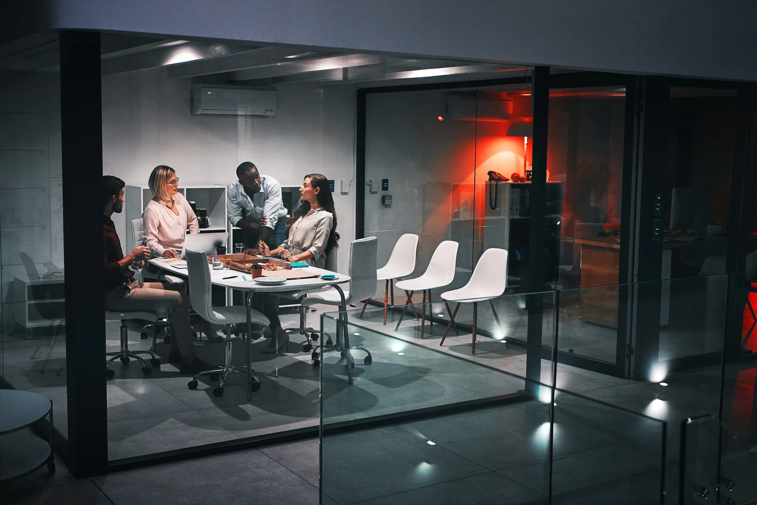 Four colleagues having a meeting around a table with laptops and pizza inside a glass-walled office room.