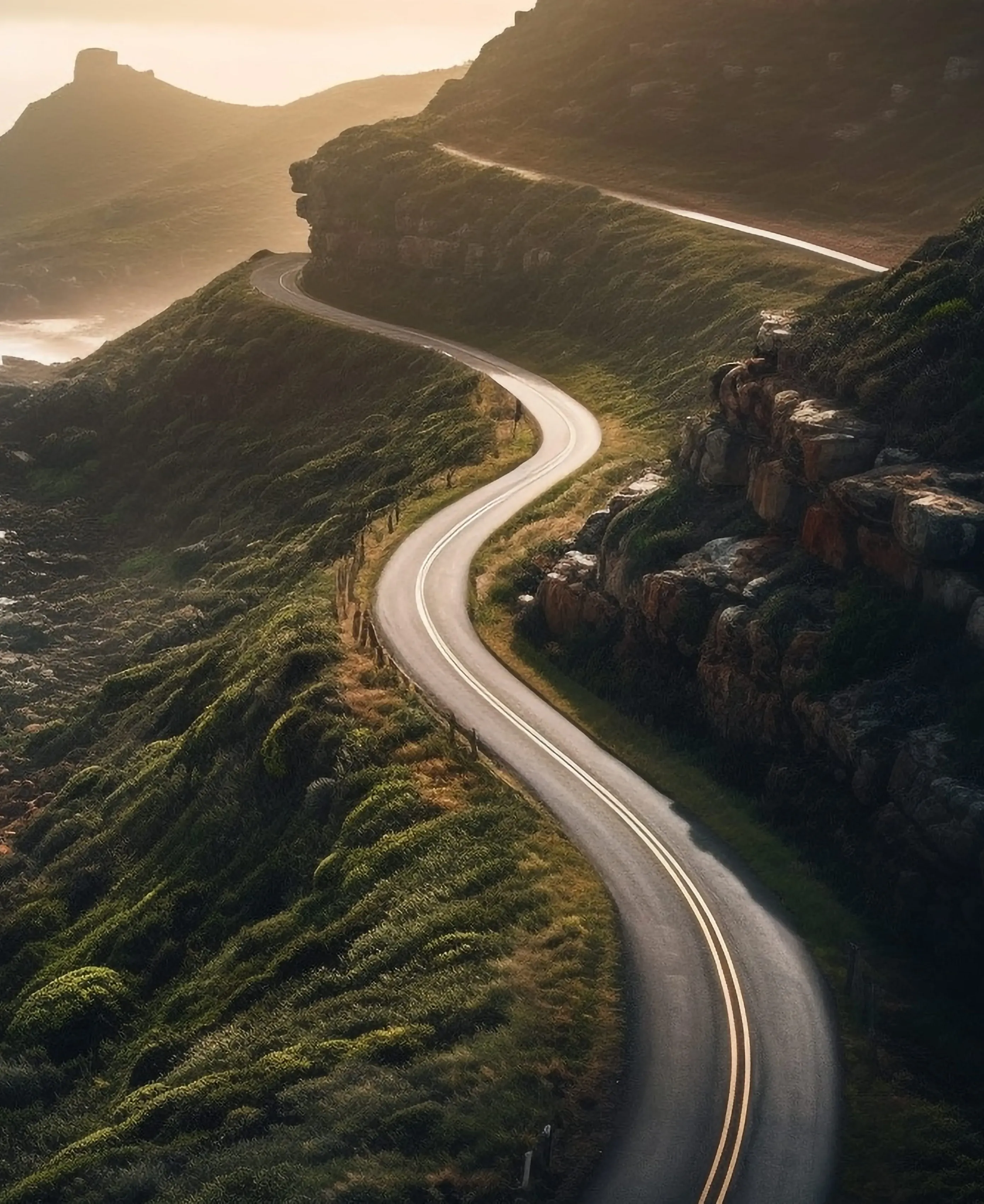Winding road curving through green hills and rocky cliffs with soft sunlight in the background.