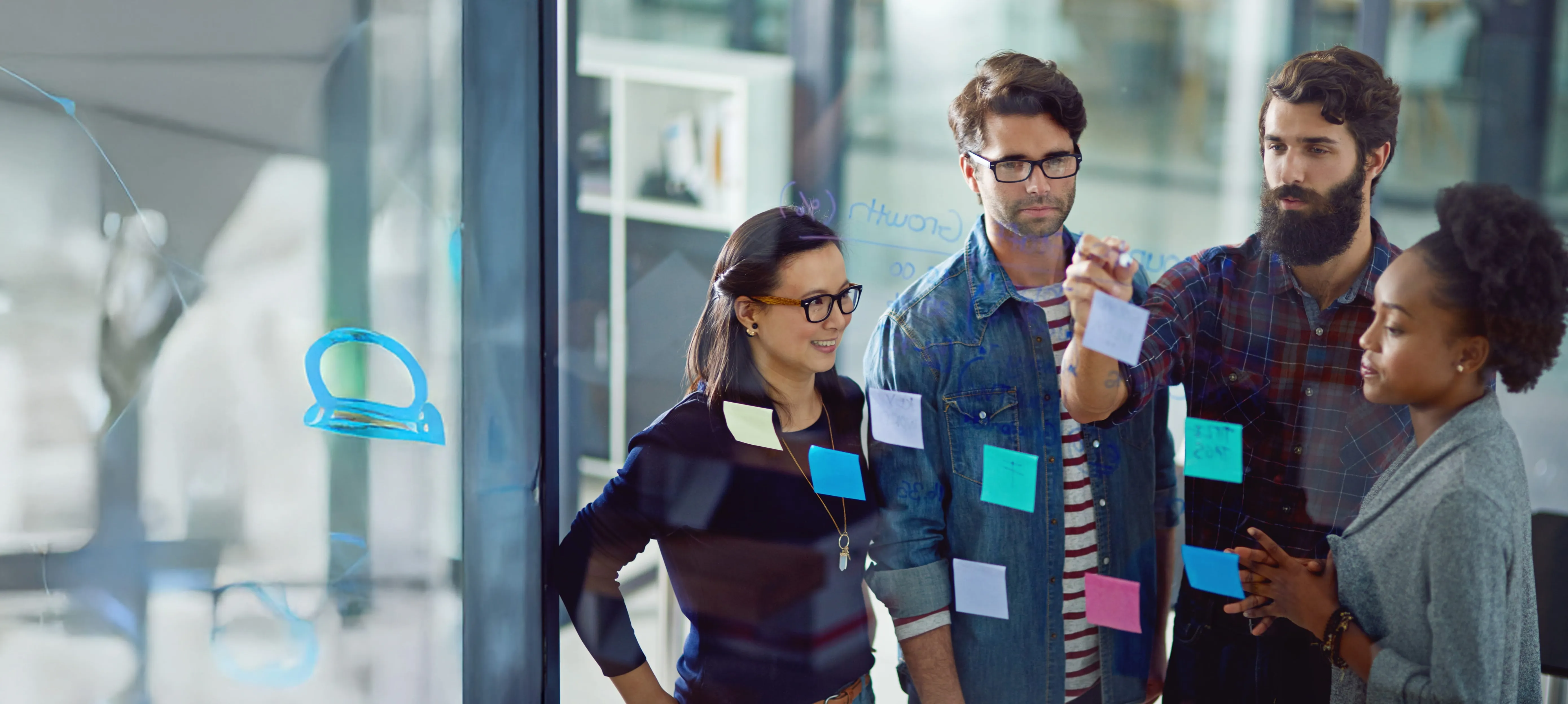 Four colleagues collaborating and discussing ideas using colorful sticky notes on a glass wall in an office.