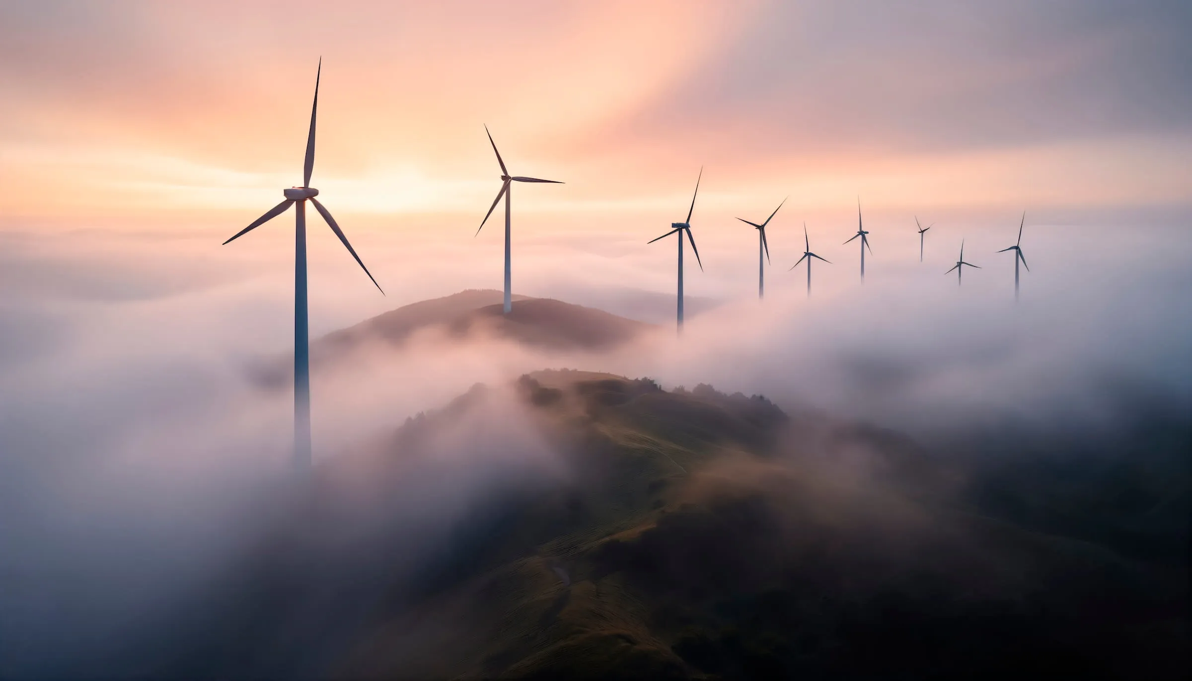 Wind turbines standing on fog-covered hills under a colorful sunrise sky.