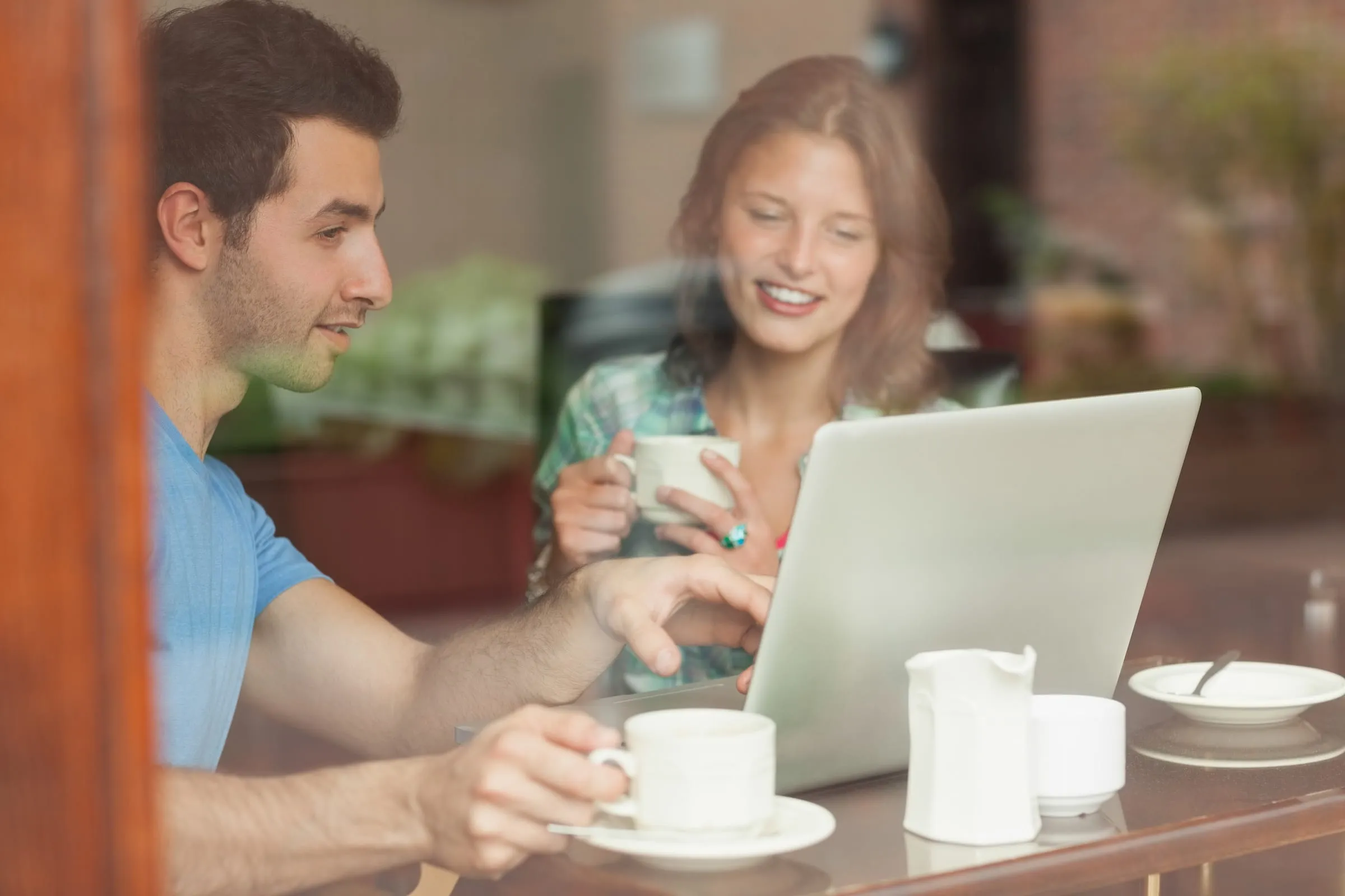 A man and woman sitting at a table with a laptop, both holding coffee cups and looking at the laptop screen.