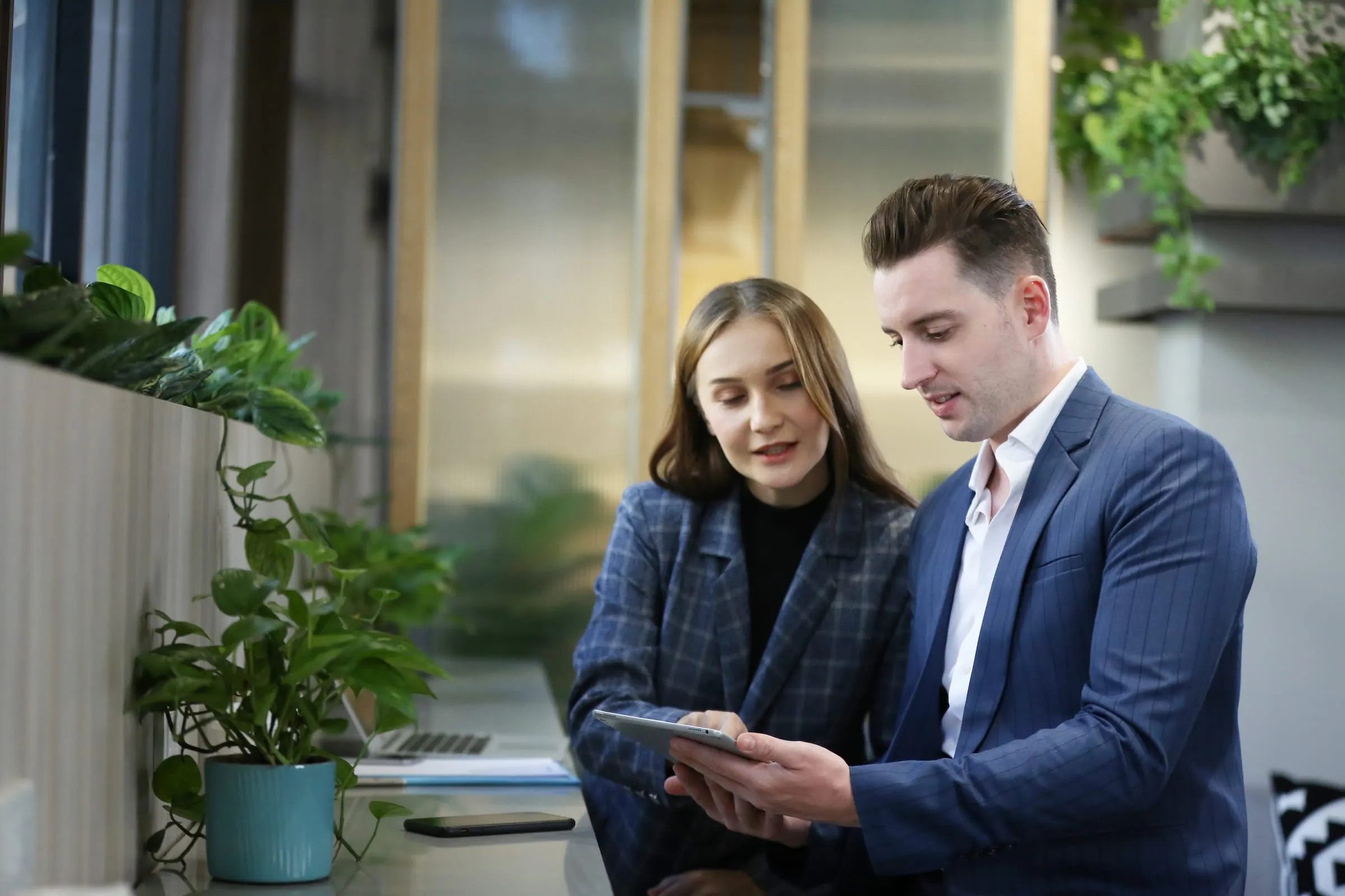 A man and woman in business attire looking at a tablet together in a modern office with plants.