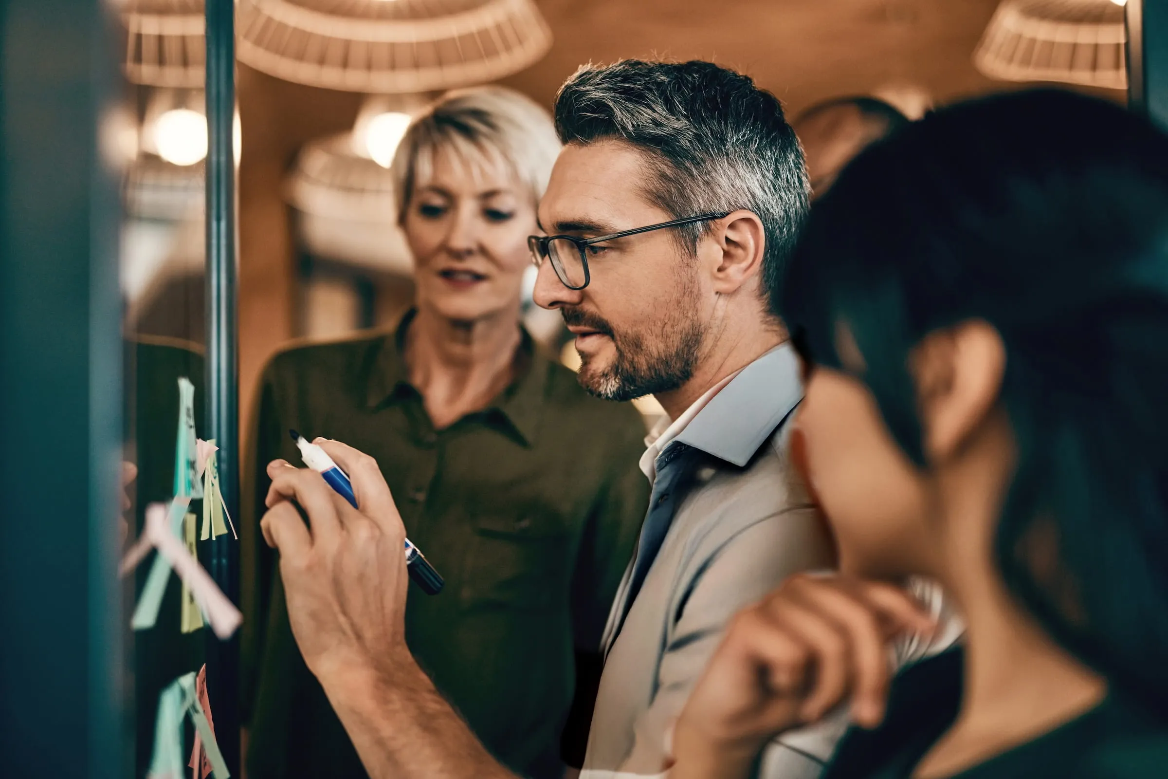 Three professionals collaborating in an office, a man writing on sticky notes on a glass wall while two colleagues watch.