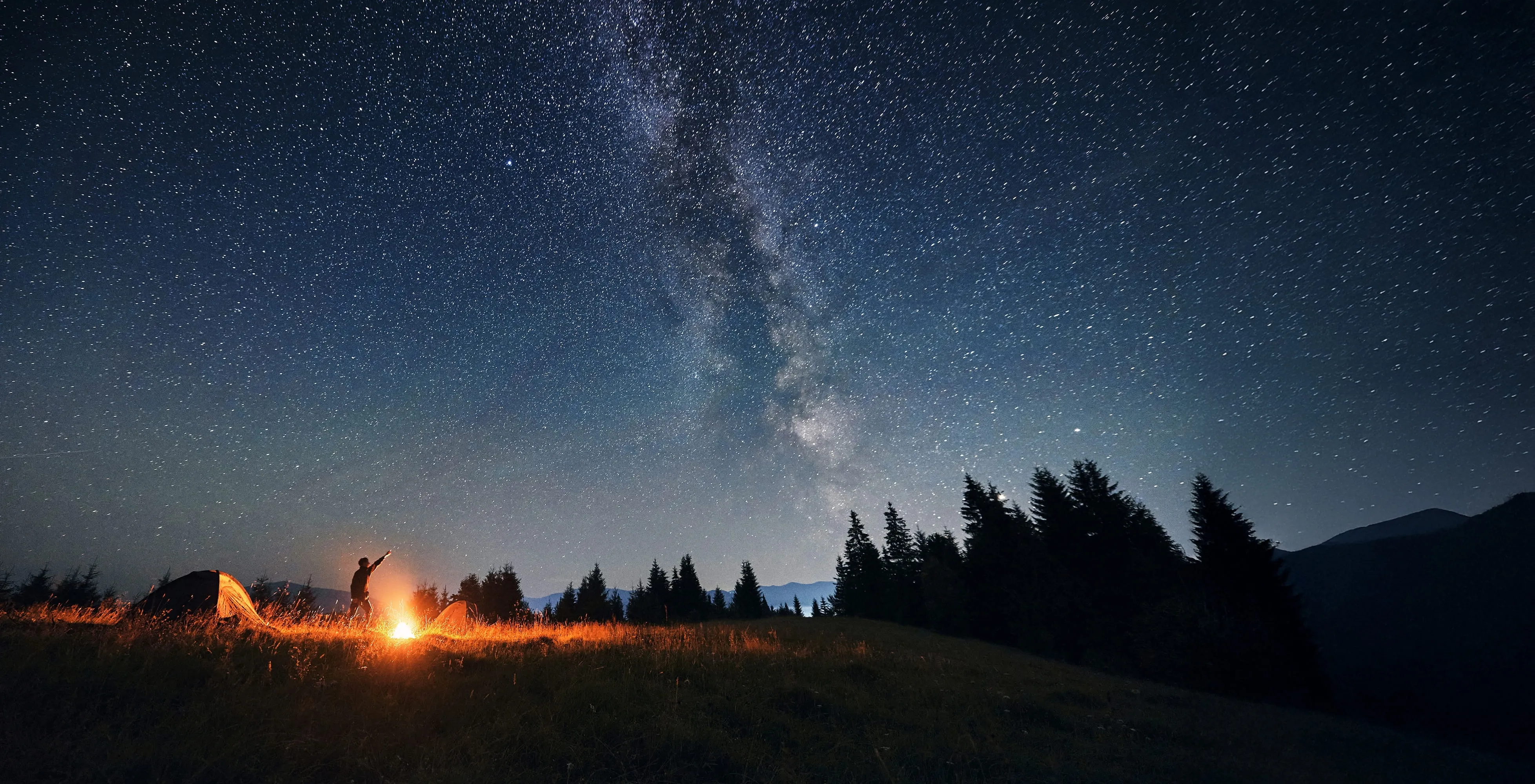 Person standing by a campfire near tents on a grassy hill under a starry night sky with the Milky Way visible.