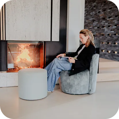 Woman sitting on a round gray chair reading a book beside a lit fireplace in a modern living room.