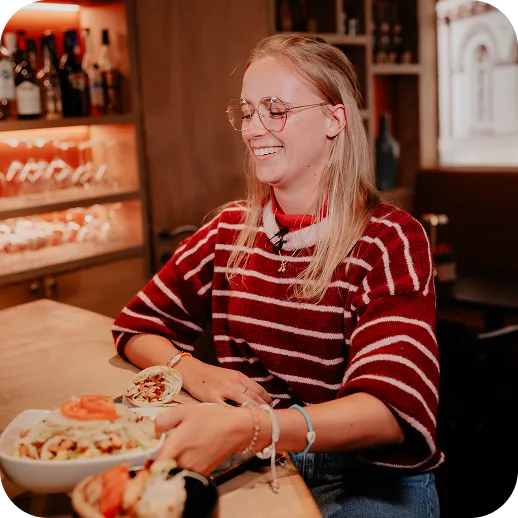 Smiling woman in glasses and a red striped sweater sitting at a table reaching for a bowl of food in a cozy restaurant setting.