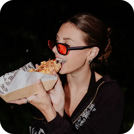 Young woman wearing red sunglasses enjoying a crispy snack in a paper tray at night.