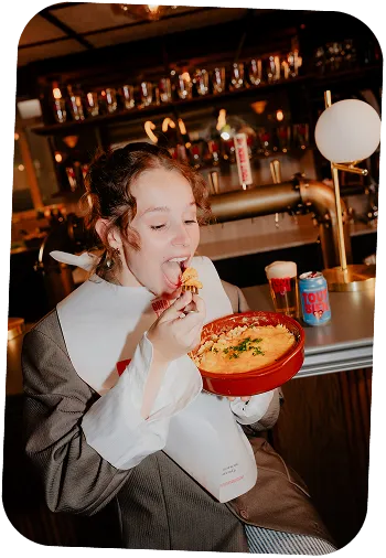 Woman wearing large white collar bib eating cheesy food from a red casserole dish inside a bar.