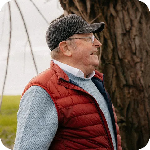 Elderly man wearing a black cap, glasses, red puffer vest, and blue sweater standing outdoors near a tree.