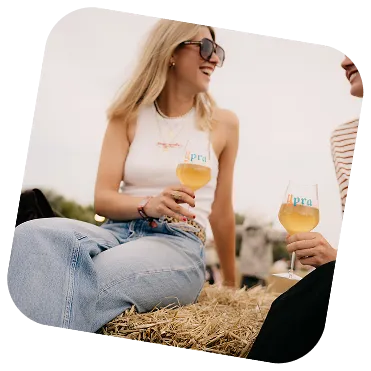 Two women sitting on hay bales outdoors, smiling and holding glasses of white wine.