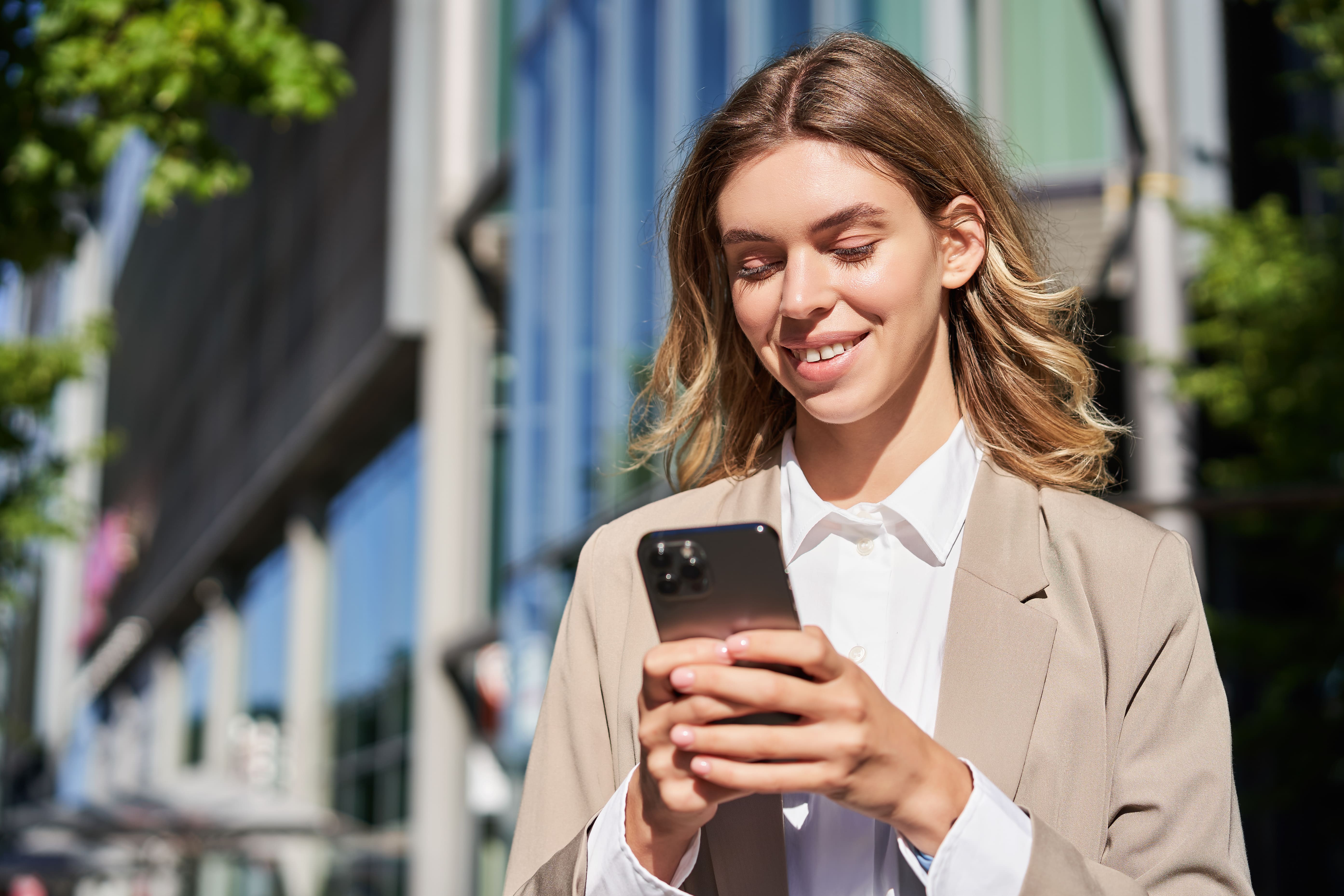 Smiling real estate advisor in a beige blazer reviewing property listings on a smartphone in front of a modern building in Tenerife, assisting international investors.
