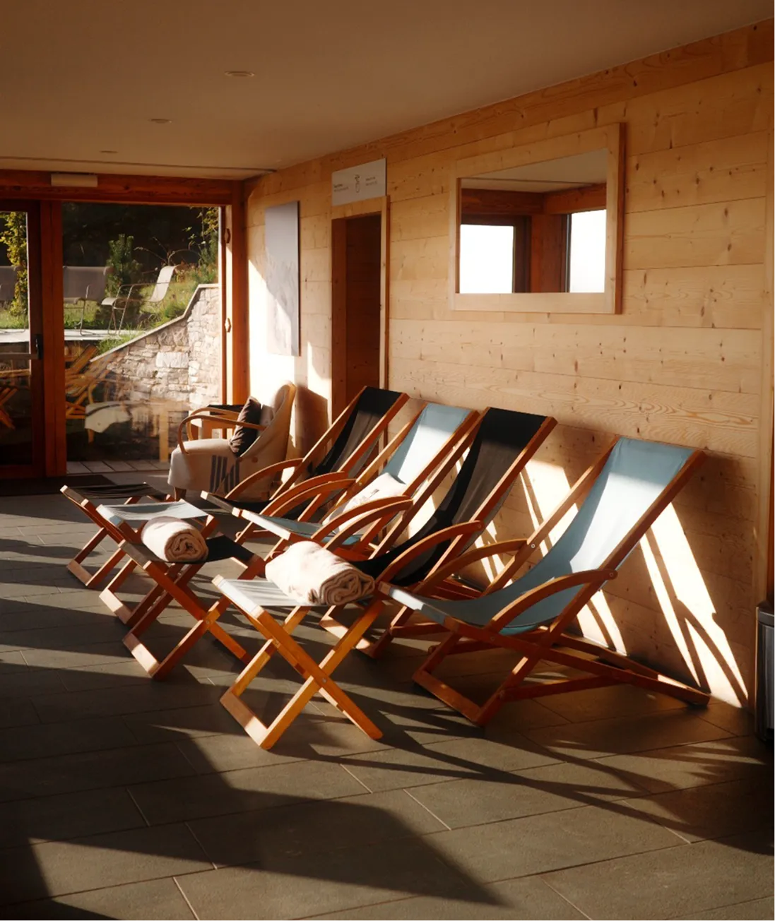 Une rangée de chaises longues en bois avec des coussins bleu clair et des serviettes roulées posées sur des petits tabourets en bois, sur un sol carrelé, avec une paroi en bois clair et une grande fenêtre reflétant la lumière du soleil.