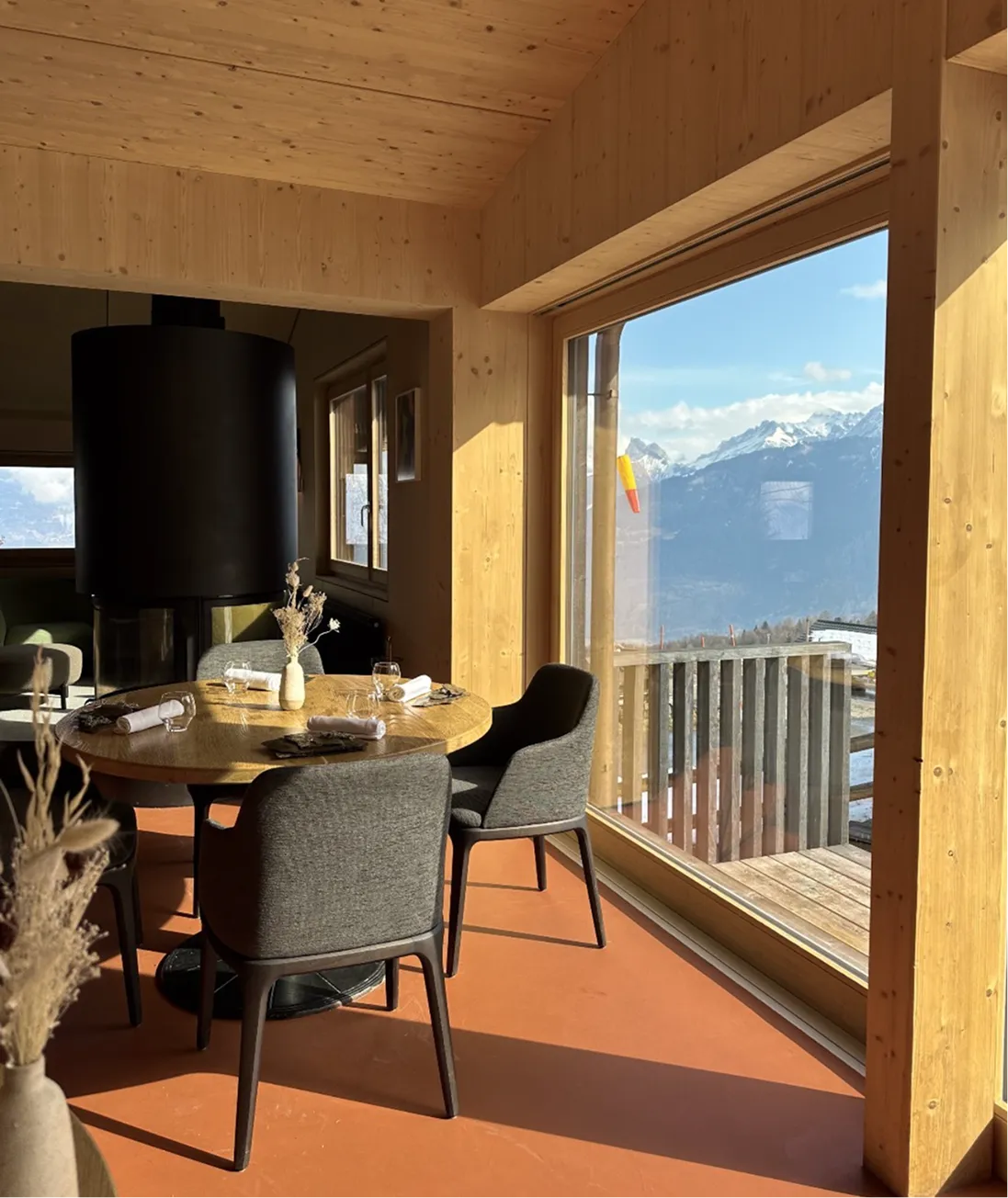 Intérieur chaleureux en bois clair avec une table ronde dressée pour quatre personnes, des chaises grises, une grande fenêtre offrant une vue sur des montagnes enneigées sous un ciel bleu et un balcon en bois.