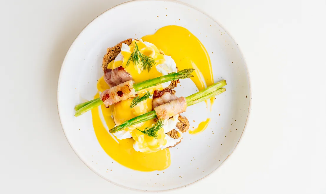 Assiette avec deux tranches de pain complet garnies d'œufs pochés nappés de sauce hollandaise, asperges enveloppées de bacon grillé, et décorées d'aneth frais.