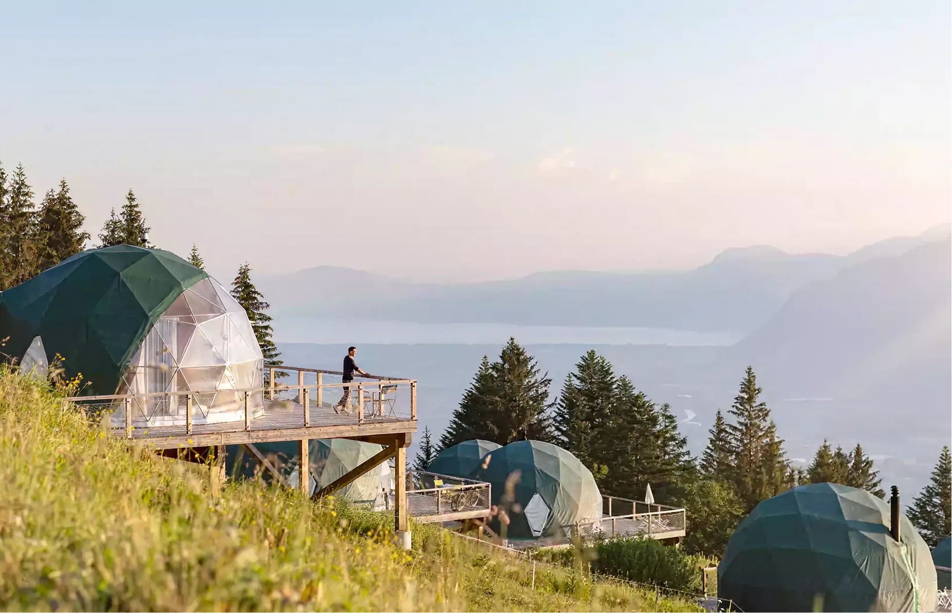 Kuppel-Pods und Holzterrassen an einem bewaldeten Hang über dem Tal – Glamping in Kuppeln im Whitepod im Wallis