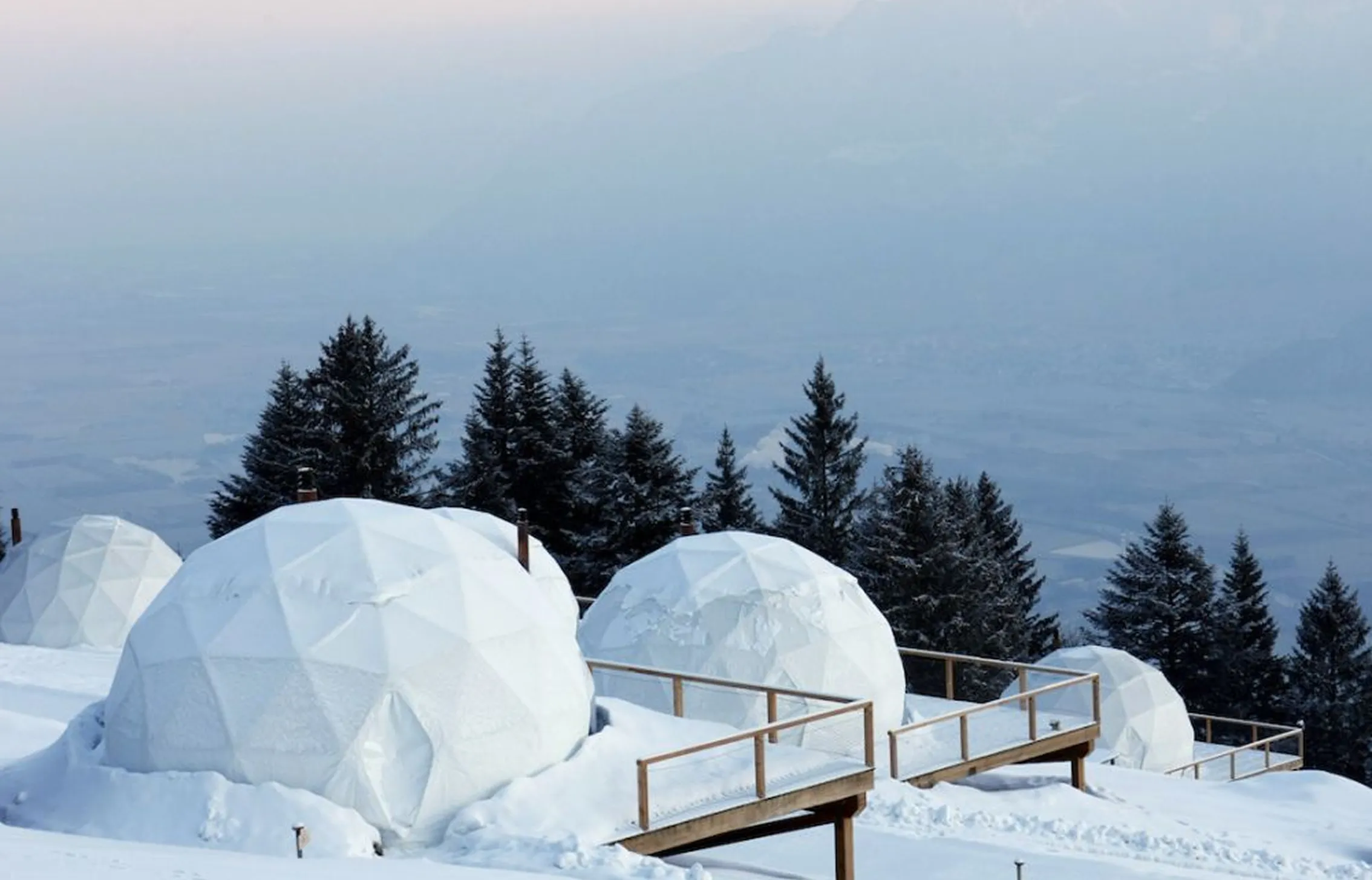 Pods en dôme enneigés au milieu des sapins dans la brume – Elle présente les igloos de Whitepod en Suisse