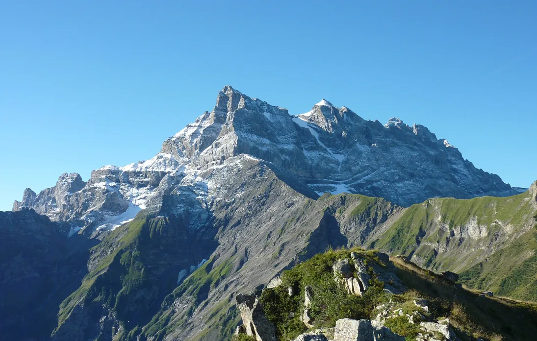 Vue sur une chaîne de montagnes avec des sommets partiellement enneigés sous un ciel bleu clair, et des pentes couvertes de végétation verte.