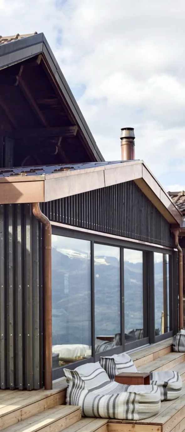 Terrasse en bois d'une maison avec grandes fenêtres vitrées reflétant des montagnes enneigées, fauteuils bas rayés posés sur les marches en bois, sous un toit à cheminée.