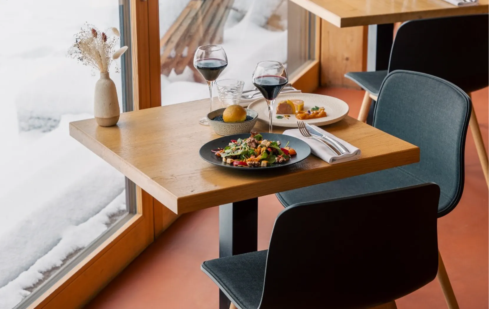 Table pour deux avec verres de vin rouge devant une baie vitrée enneigée – restaurant Les Cerniers au-dessus de Monthey