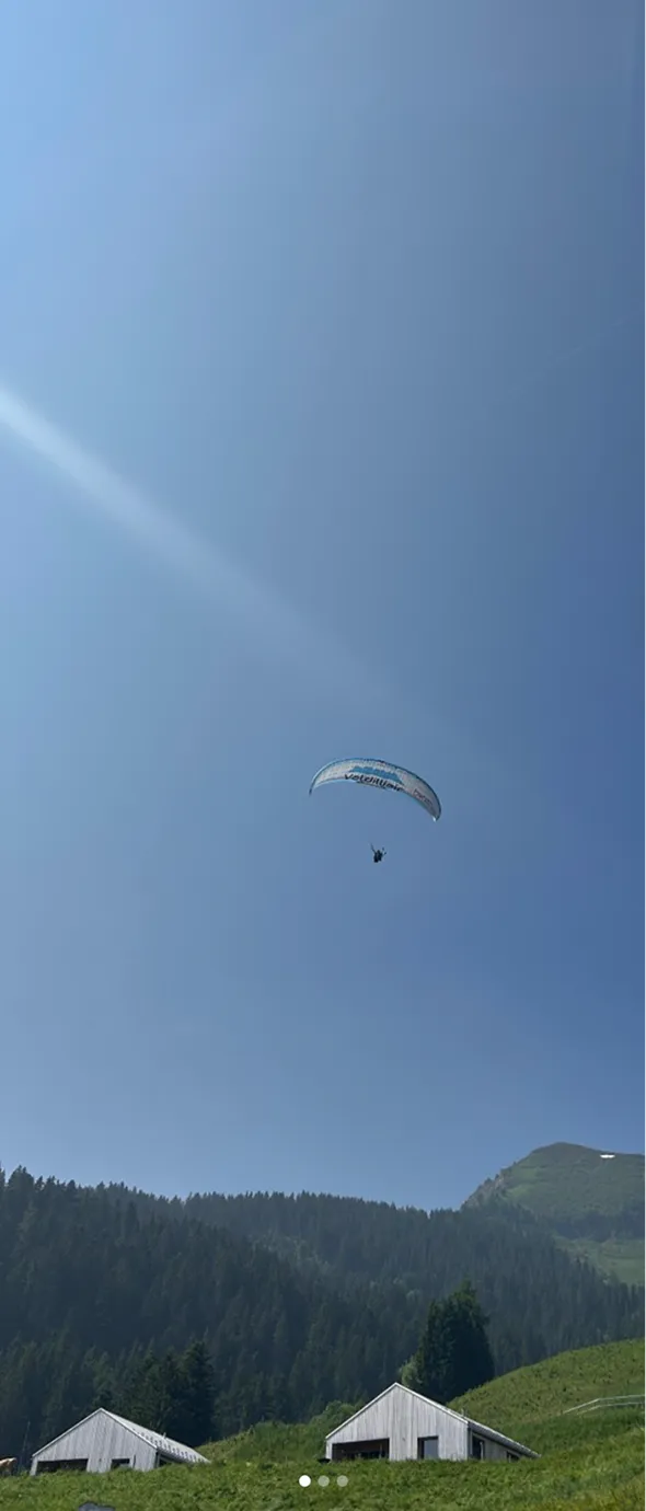 Parapentiste volant sous un ciel bleu clair au-dessus d'une colline verte avec deux maisons blanches en contrebas et une forêt dense en arrière-plan.