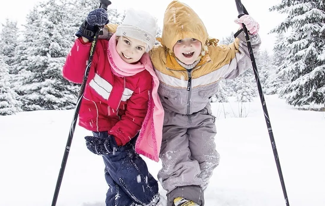 Deux enfants habillés chaudement en tenues de ski colorées, souriants, tenant des bâtons de ski dans la neige avec des arbres enneigés en arrière-plan.