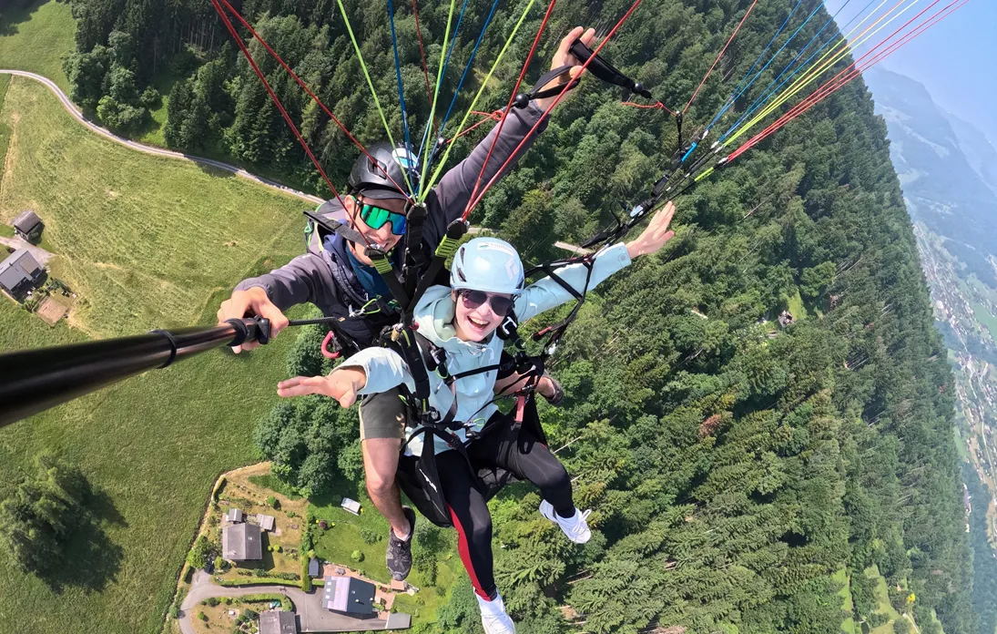 Deux personnes faisant un parapente en tandem au-dessus d'une forêt et de maisons isolées, avec un homme tenant un selfie stick pour prendre la photo.
