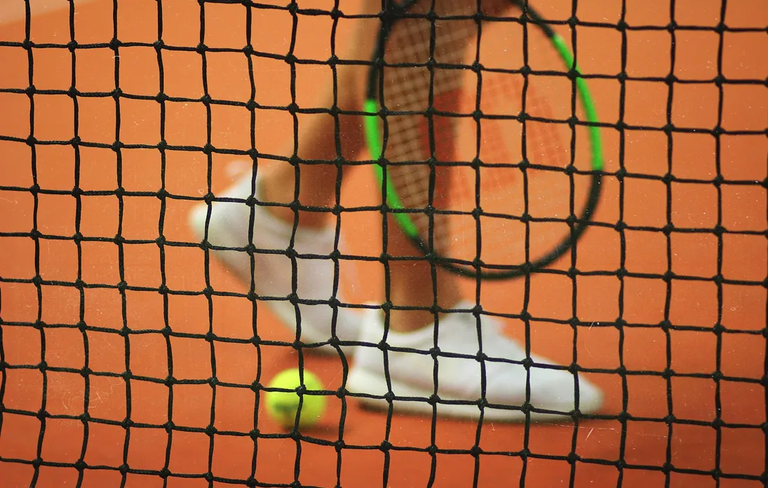 Vue à travers un filet de tennis sur un terrain en terre battue montrant un joueur avec des chaussures blanches, une raquette avec un cadre vert, et une balle jaune au sol.