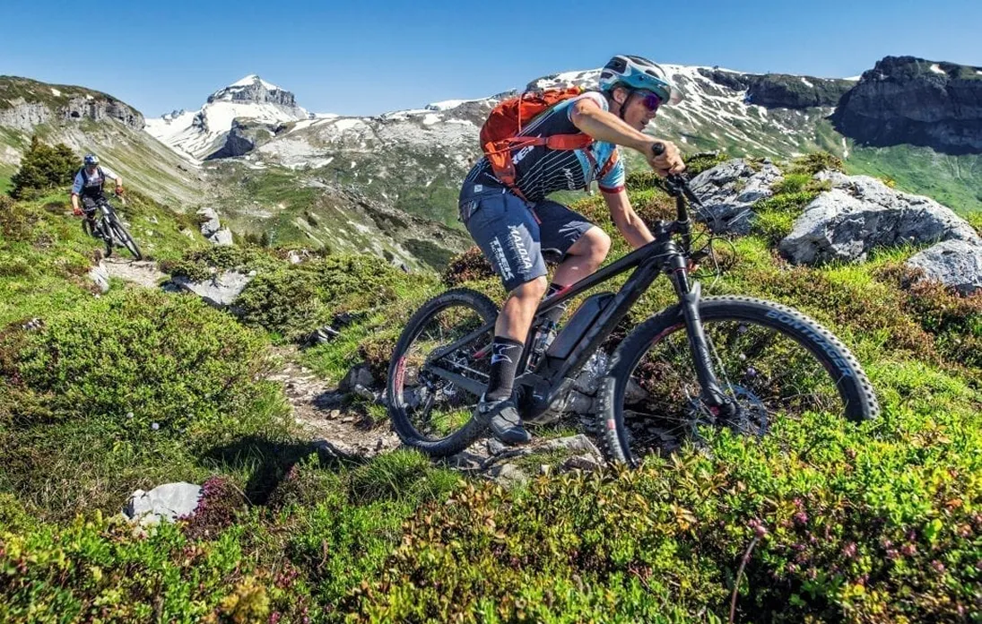 Deux vététistes descendent un sentier étroit en montagne, entourés de rochers et de végétation avec des sommets enneigés en arrière-plan sous un ciel bleu clair.