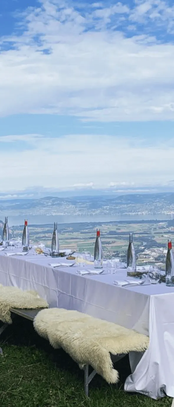 Table extérieure longue dressée avec nappe blanche, verres, couverts, bouteilles d'eau avec bouchons rouges, et bancs recouverts de plaids en laine, avec vue panoramique sur paysage vallonné et ciel partiellement nuageux.