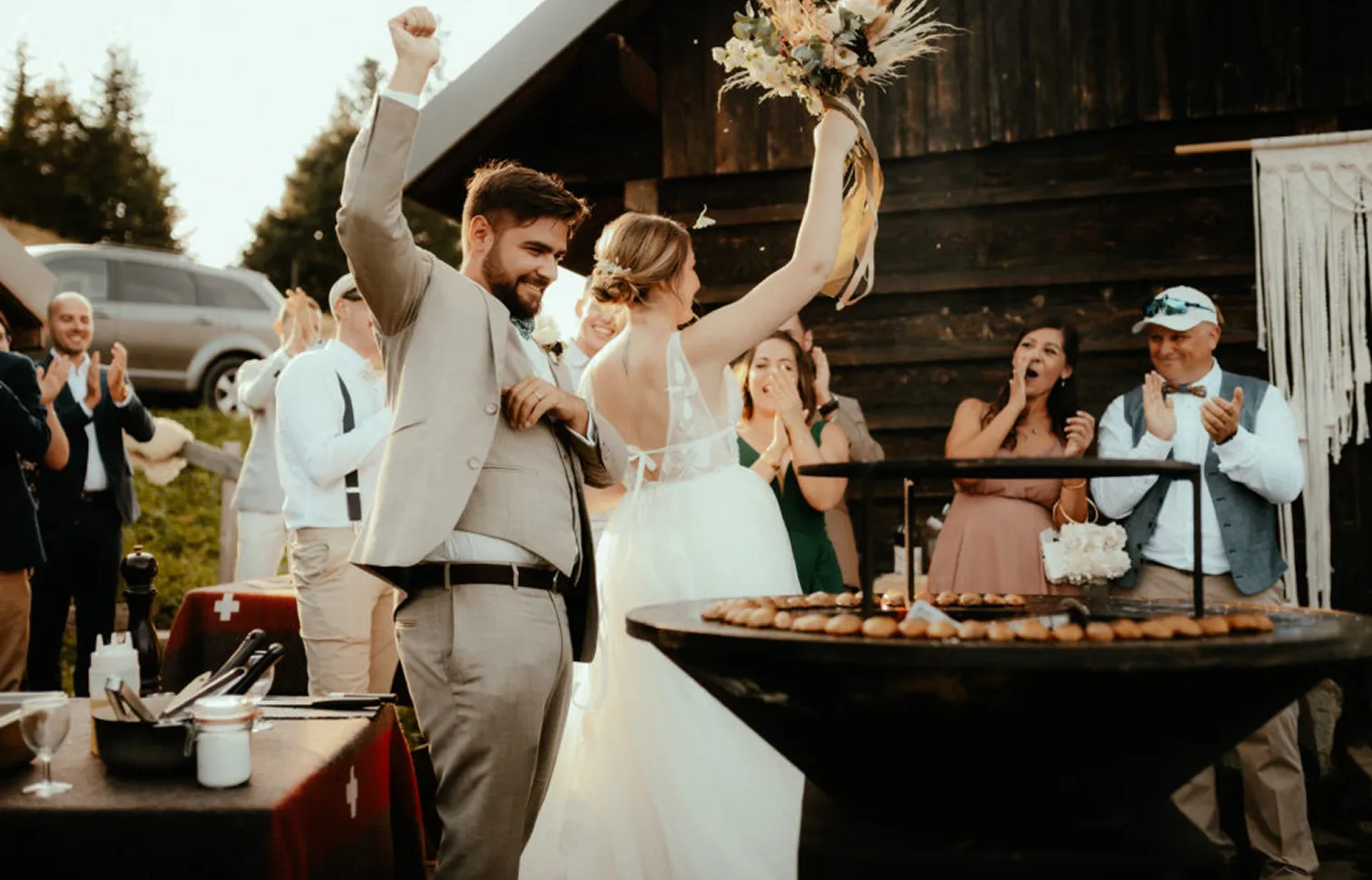 Un couple de mariés célèbre avec joie, la mariée lève un bouquet de fleurs, entourés de leurs invités qui applaudissent lors d'une réception en plein air près d'une maison en bois.