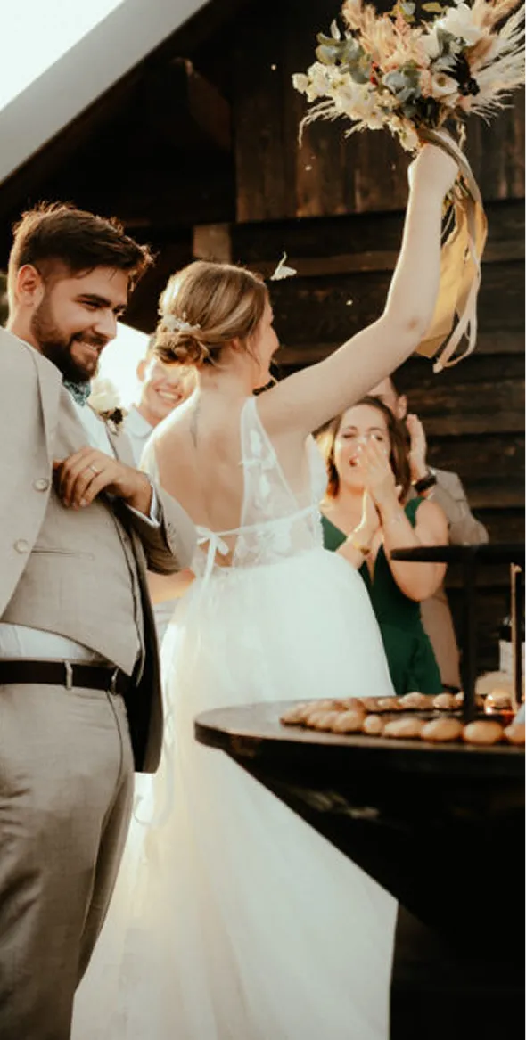 Un couple de mariés souriants, la mariée de dos brandissant un bouquet de fleurs séchées, entouré d'invités applaudissant lors d'une réception en plein air.