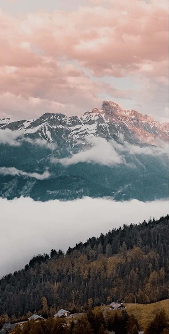 Montagnes enneigées avec un ciel rose au coucher du soleil, des nuages flottant au-dessus et une forêt dense en bas avec quelques maisons éparses.