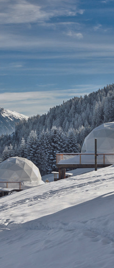 Deux dômes géodésiques blancs installés sur une terrasse en bois sur une pente enneigée, avec une forêt de conifères enneigés et des montagnes en arrière-plan sous un ciel partiellement nuageux.