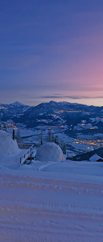 Vue nocturne d'un paysage enneigé avec deux igloos au premier plan et des montagnes illuminées par les lumières d'un village au loin sous un ciel bleu et violet clair.