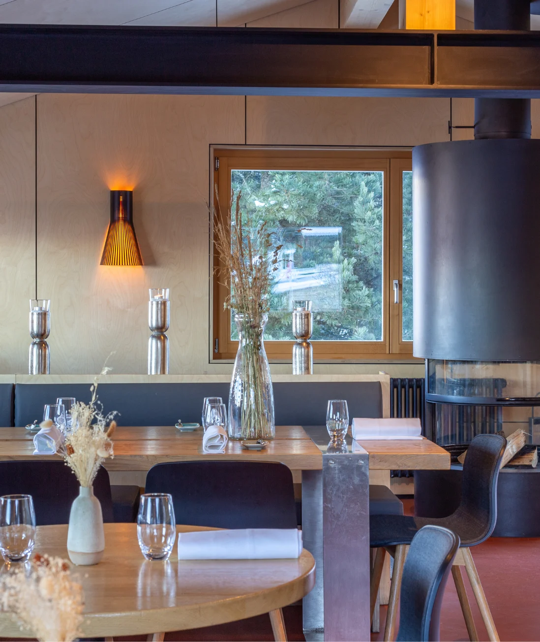 Intérieur d'un restaurant moderne avec tables en bois décorées de vases contenant des plantes séchées, chaises noires, un grand poêle noir et une fenêtre montrant des arbres verts à l'extérieur.