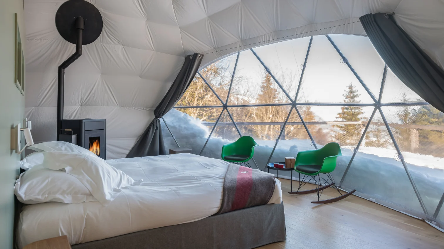 Bett, schwarzer Ofen und grüne Sessel vor dem verschneiten Pod-Fenster – Familien Pod, Pod von Whitepod im Wallis