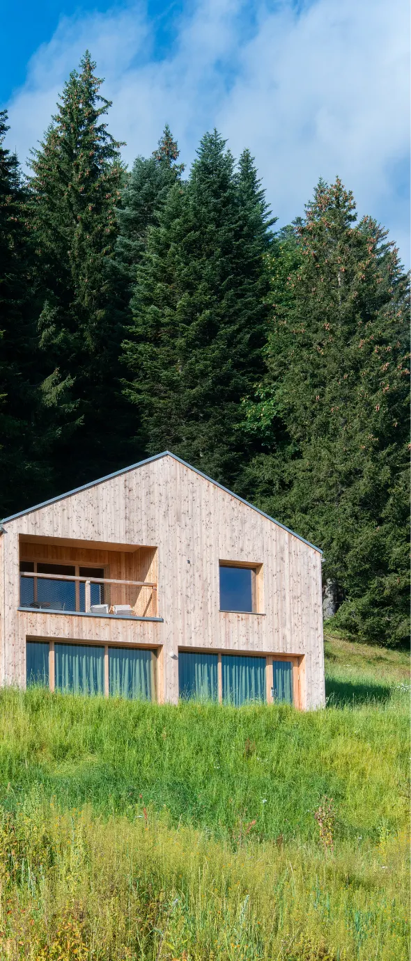 Maison en bois clair avec grande fenêtre au rez-de-chaussée et balcon à l'étage, située dans une prairie verte devant un groupe dense de grands sapins sous un ciel partiellement nuageux.
