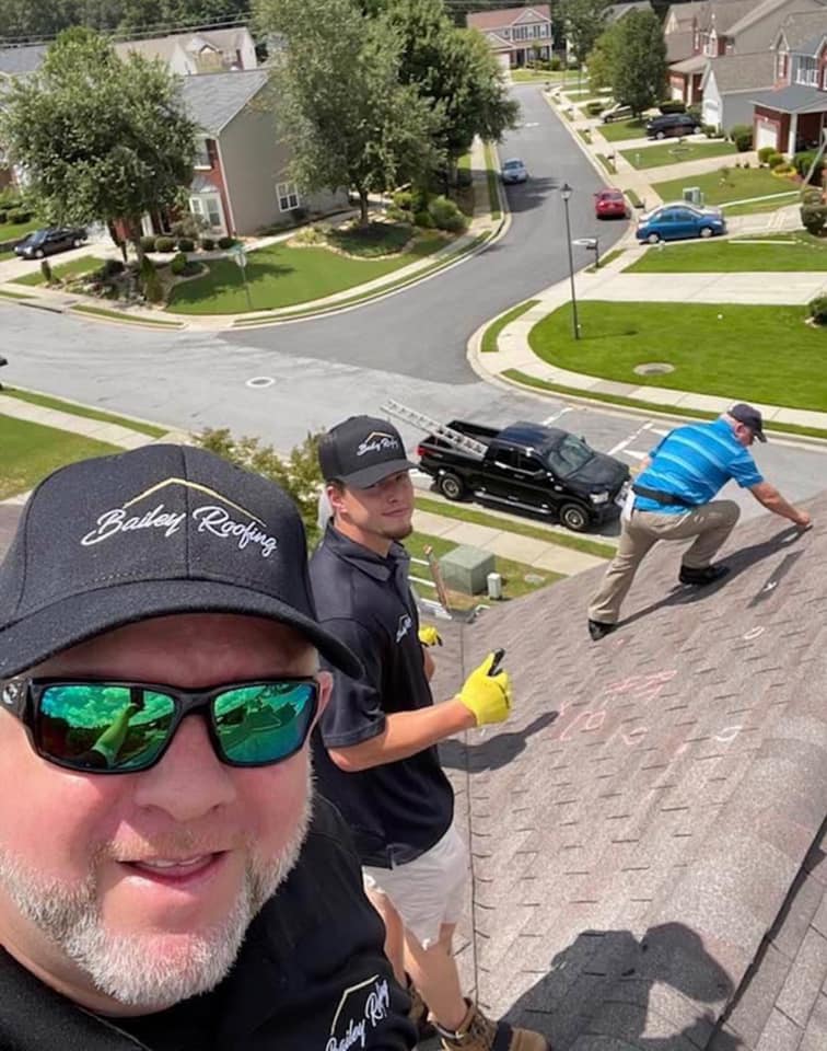 Three roofing workers on a residential roof, one taking a selfie wearing Bailey Roofing cap and sunglasses, another giving a thumbs up, and the third bending to work on the shingles.